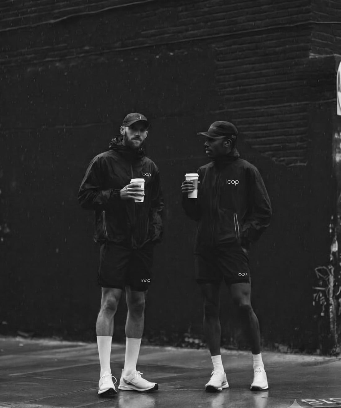 Two men in black Loop rain jackets and shorts holding coffee on a wet street
